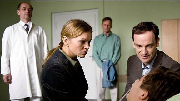 Vier Personen stehen um das Krankenbett eines jungen Mannes herum; sie sehen besorgt aus. 