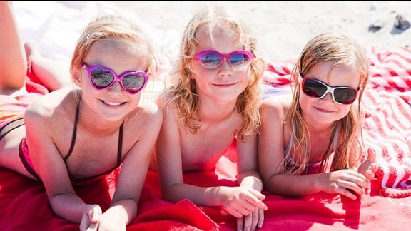 Ferienlager - drei blonde Mädchen mit Sonnenbrillen liegen auf einer Decke am Strand
