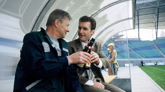 Georg Bracht (Günter Junghans, links) und sein Sohn Daniel Bracht (Siegfried Terpoorten) sitzen auf einer Stadionbank und stoßen mit Bier an.