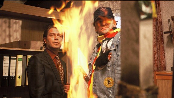 ost Meckel (Zacharias Preen) Dirk Jamas (Michael Günther, rechts) stehen in einem Raum neben einer großen Flamme.
