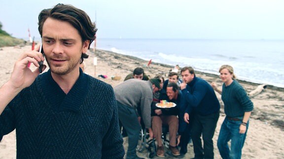 Im Vordergrund telefoniert ein Mann, im Hintergrund hat sich eine Gruppe am Strand um eine Geburtstagstorte versammelt.