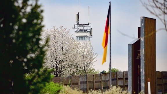 Ein Denkmal mit deutscher Fahne steht vor einer Mauer; im Hintergrund ragt ein ehemaliger Wachturm in den Himmel