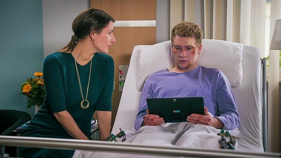 Frau sitzt am Krankenbett eines liegenden und verletzten Mannes, der in einen Laptop schaut
