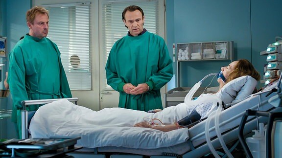 zwei Ärzte stehen vor dem Bett einer frisch operierten Patientin mit Atemmaske