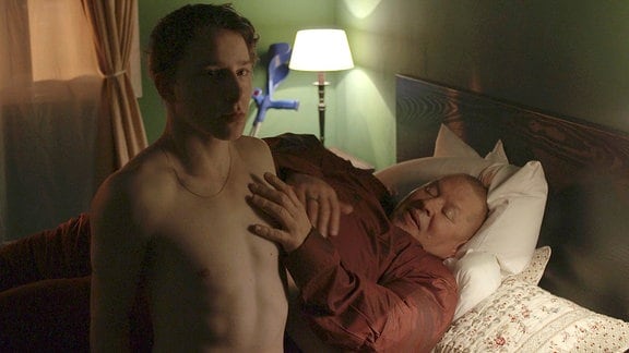 Felix steht nackt am Bett eines dicken Mannes.
