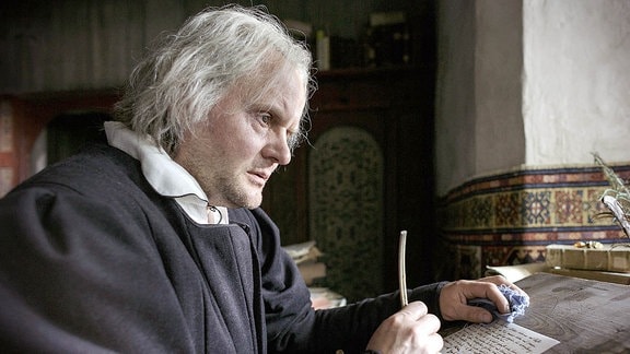 Ein Mann in einer mittelalterlichen Schreibstube nachdenklich mit Schreibfeder in der Hand.