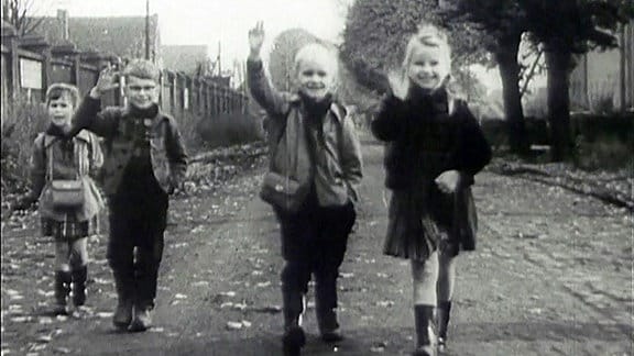 Vier Kinder stehen auf einer Straße und winken in die Kamera.