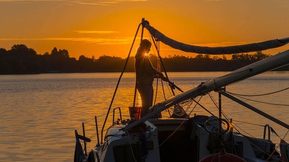 Silhouette einer Frau auf einem Segelboot bei Sonnenuntergang mit orangefarbenem Himmel.