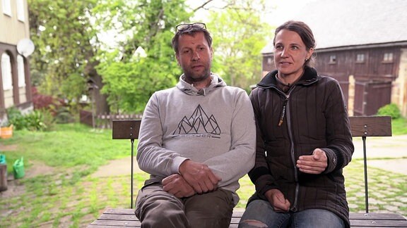 Ein Mann und eine Frau in einem ländlichen Hof sitzen nebeneinander auf einer Bank.