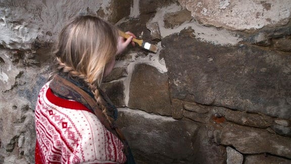 Eine junge Frau mit geflochtenen Zöpfen streicht mit einem Pinsel an den Fugen einer Bruchsteinmauer.