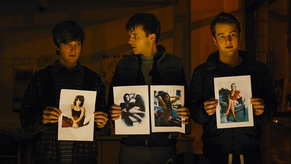Drei junge Männer stehen nebeneinander und halten Fotos mit leichtbekleideten Frauen in den Händen.  