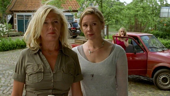 Eine ältere Frau steht neben einer jüngeren Frau. Dahinter steht eine weitere, junge Frau an einem Auto.