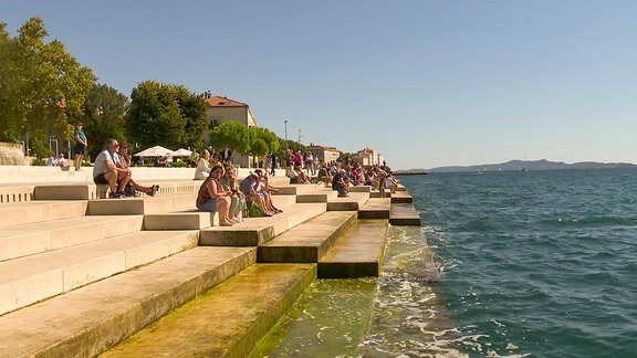 Blick auf ein Ufer mit breiteen Treppen, auf denen viele Menschen sitzen.