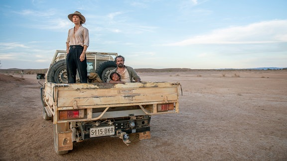 Eine stehede Frau mit Hut und ein sitzender Mann mit einem Jungen im Arm auf der Ladefläche eines Pickup, der in einer kargen Landschaft steht.