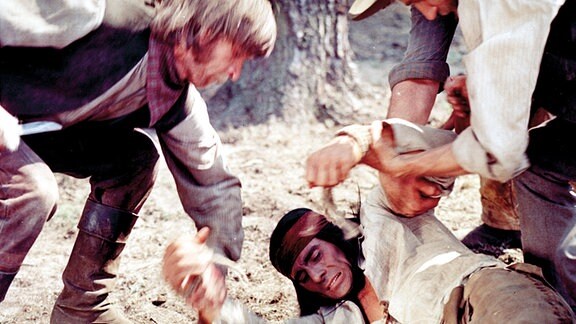 Der junge Mimbreno-Häuptling Ulzana (Gojko Mitic, Mitte liegend) ist in eine Falle der weißen Trapper geraten, die zu korrupten Mineningenieur Johnson gehören. Der übt Rache an dem jungen Häuptling und lässt ihn an einem Baum fesseln und auspeitschen.