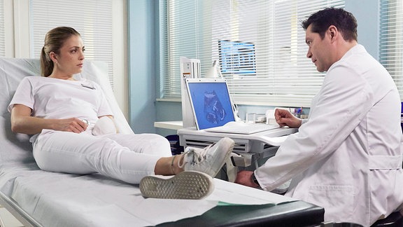 Eine Krankenschwester sitzt auf einer Liege. Sie sieht einen Arzt an, der neben ihr an einem Bildschirm sitzt.