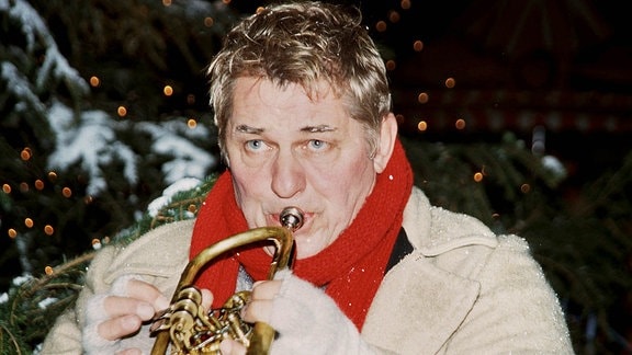 Johannes spielt mit ernstem Blick Trompete. Er trägt Winterkleidung, im Hintergrund ein Weihnachtsbaum. 