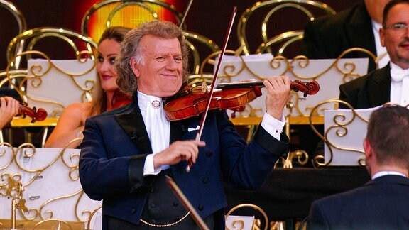 Ein lächelnder Mann spielt Geige vor einem Orchester.