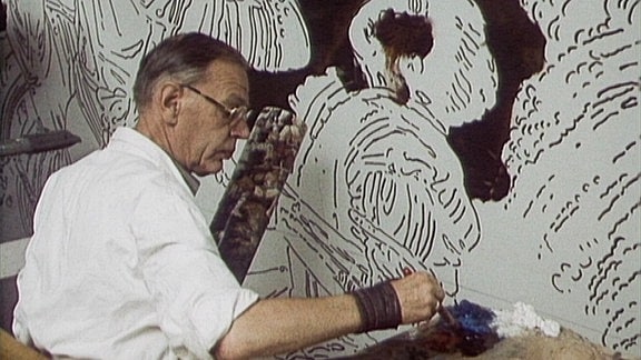 Ein Maler steht vor seinem Bild und nimmt mit dem Pinsel Farbe von einer Palette auf.
