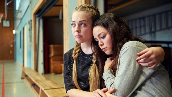 Zwei junge Frauen sitzen auf einer Bank in einer Turnhalle. Die eine hat den Arm tröstend um die Schulter der anderen gelegt. Beide schauen besorgt und traurig.