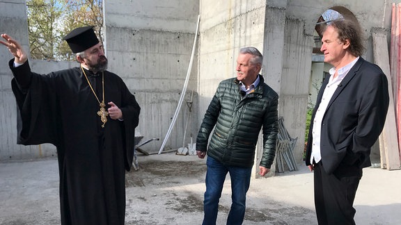 Eine Person im Gewand eines christlichen griechisch-orthodoxen Priesters spricht mit zwei anderen Personen. Im Hintergrund eine Kirchen-Baustelle.