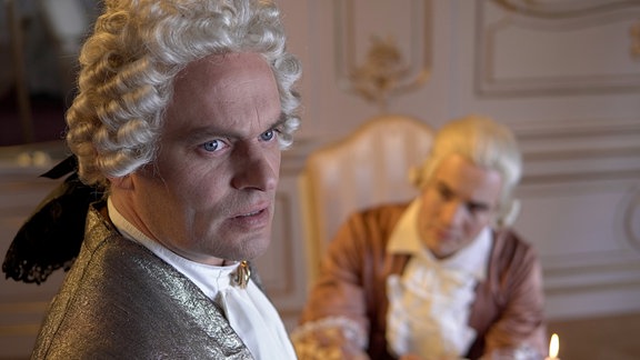Ein Mann in der Kleidung des 18. Jahrhunderts schaut erregt in die Ferne, ein anderer Mann, der neben ihn sitzt, betrachtet ihn.