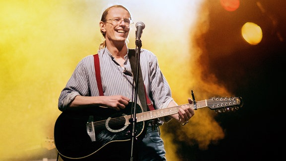 Ein Mann mittleren Alters steht mit einer Gitarre auf einer erleuchteten Bühne.