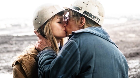 Ein junger Mann mit Bauarbeiterheld küsst eine junge Frau mit Bauarbeiterhelm.