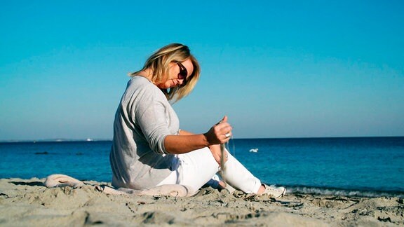 Jana am Strand der Ostsee sitzend. Ihre rechte Hand lässt Sand aus dieser rieseln.