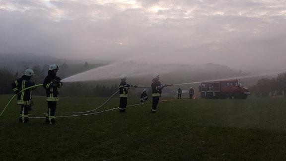 Mehrere Personen in Feuerwehr-Kleidung stehen auf einer nebligen Wiese und halten Löschschläuche, aus denen Wasser spritzt. Im Hintergrund ein Feuerwehrauto.
