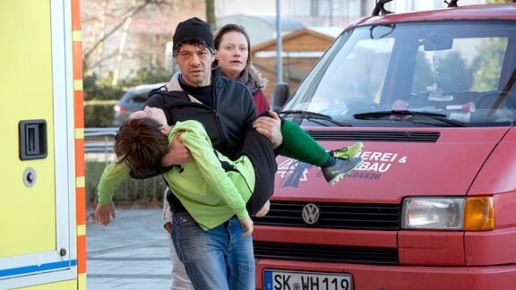 Frank Baumann (Carlo Ljubek) trägt Hannes Baumann (Mikke Rasch) in die Notaufnahme, gefolgt von seiner Ex-Frau Esther Baumann (Katharina Marie Schubert)