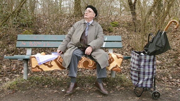 Ein alter Mann sitzt auf einer Parkbank mit ausgerollter Decke, etwas Essen und einem Handwagen. Im Hintergrund sieht man einen winterlichen Wald.