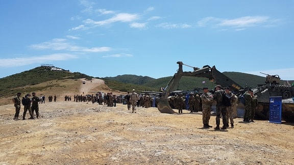 Auf einem Berg stehen auf einer offenen Fläche viele Soldaten und Panzer. Bei sonnigem Wetter ist die Lage entspannt.