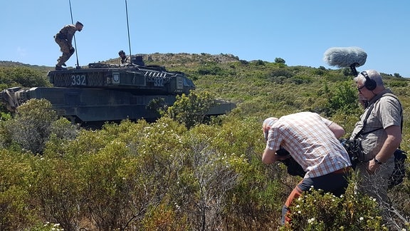 In einer hügeligen Landschaft fährt duch eine mit Büschen bewachsenen Szenerie ein Panzer, ein Kamerateam filmt diesen.