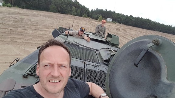 Auf einem von Wald umgebenen Feld steht ein Panzer, aus dessen drei Luken Menschen mit ziviler Kleidung in die Kamera schauen.