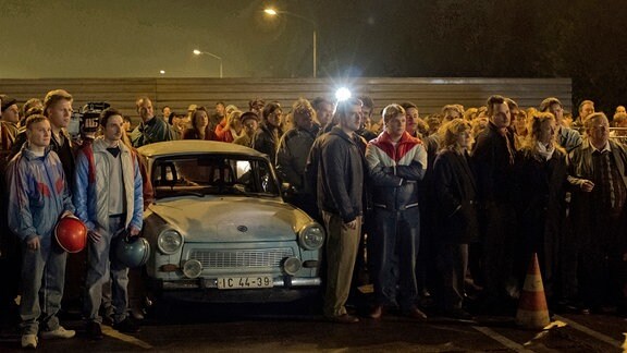 Eine Menschengtuppe - in ihr ein "Trabant 601" - steht wartend vor einem Metallzaun. Eine Fotolampe erhellt die dunkle Umgebung.
