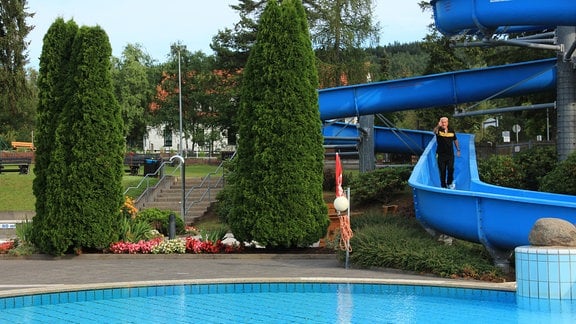 Eine blaue, kurvige Wasserrutsche führt in ein Freibad-Schwimmbecken. Ein Mann steht bekleidet auf der Rutsche und streckt seinen Daumen nach oben.