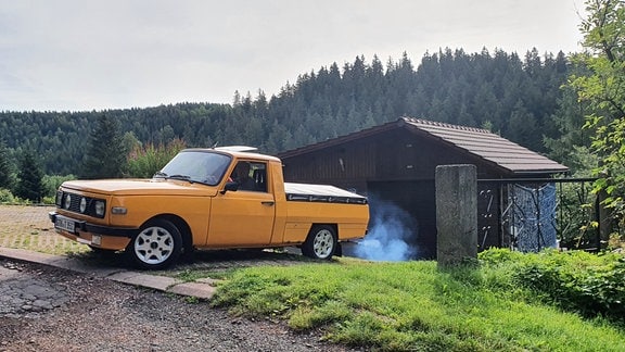 Ein gelber Pick-up Wartburg auf einer Einfahrt. Im Hintergrund eine Holzhütte und Wald.