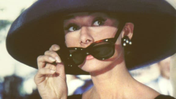 Audrey Hepburn als Holly Golightly setzt ihre große Sonnenbrille ab. Sie trägt einen großen Hut.
