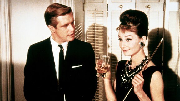 Audrey Hepburn als Holly Golightly hat ein Glas und eine Zigarettenspitze in der Hand und schaut erfreut. Neben ihr steht George Peppard als Paul Varjak und sagt etwas zu ihr.