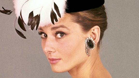 Portraitfoto von Audrey Hepburn als Holly Golightly, sie trägt ein schwarzes Kleid, große schwarze Ohrringe und einen schwarzen Hut mit auffälliger Verzierung