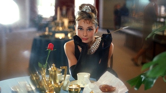 Audrey Hepburn als Holly Golightly sitzt in eleganter Kleidung und mit einer Zigarettenspitze in der Hand an einem Tisch in einem Café, vor ihr goldfarbenes Geschirr.