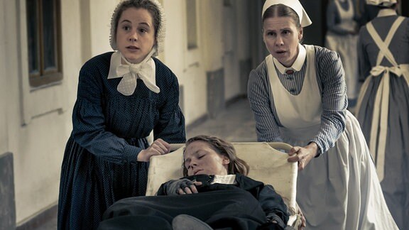 Ida Lenze (Alicia von Rittberg, M.) wird mit Verdacht auf Blinddarmentzündung in die Charité eingeliefert. Schwester Therese (Klara Deutschmann, l.) und Wärterin Edith (Tanja Schleiff, r.) verlieren keine Zeit.