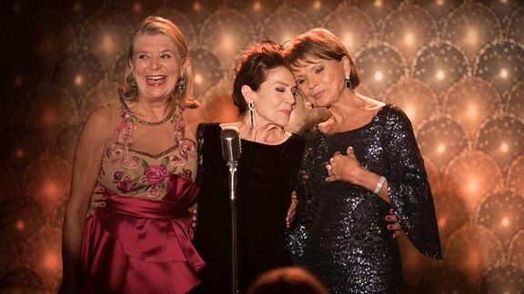 Drei ältere Frauen auf einer Bühne stehen hinter einem Mikrofon.
