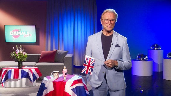 Ein Mann im Anzug steht in einem Studio und hält ein Fähnchen mit englischer Flagge in der Hand.