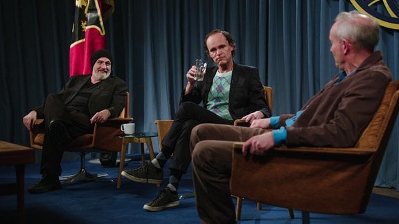 Drei Männer sitzen in Sesseln auf einer Bühne. Einer erhebt ein Glas.