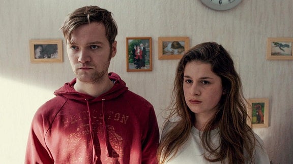 Ein junger Mann und eine junge Frau stehen in einem Wohnraum mit kleinen Bildern an der Wand.