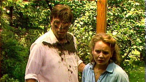 Ein Mann mit dreckverschmiertem Gesicht und eine Frau in einem Waldstück schauen entsetzt.