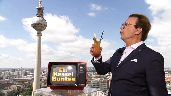 Ein Mann mit Cocktailglas steht nebe einem kleinen Fernseher vor einem Bild von Berlin.