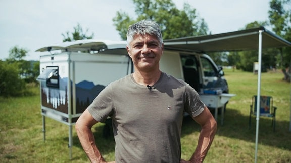 Ein Mann steht lächelnd vor einem Campingfahrzeug.